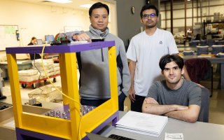 Three young men appear with a gold and purple device with lots of buttons on it. One of the men is seated folding his arms. The device has a large button on top.