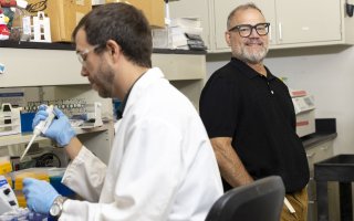 In the foreground, a man wearing a white lab coat and clear protective glasses handles a pipette. To the right, standing behind him is a man wearing a black polo shirt and black square rimmed glasses. 