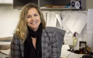 A smiling blond woman in a gray checked jacket and black scarf sits at a cluttered desk.