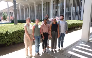 Six people, including four women and two men, stand together, smiling for a group portrait. They are standing outside, sheltered by the roof of UAlbany's academic podium. A row of evergreen bushes and academic buildings can be seen in the background.