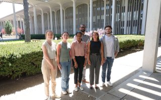 Six people, including four women and two men, stand together, smiling for a group portrait. They are standing outside, sheltered by the roof of UAlbany's academic podium. A row of evergreen bushes and academic buildings can be seen in the background.