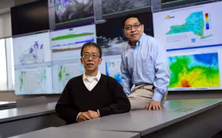 Professors Aiguo Dai and Liming Zhou sit at a table in front of weather maps at ETEC.