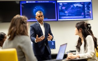 A man in a blue suit stands in front of computer screens gesturing in front of his body while talking to two students.