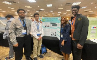 Four students — three boys and a girl wearing name tags and dress clothes — stand next to a poster board depicting air pollution research in a hotel conference room.