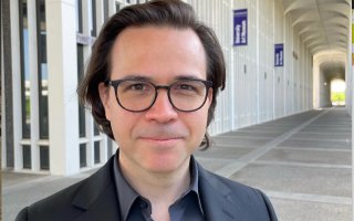 A man with short brown hair and black-rimmed glasses smiles outside of the University Art Museum at UAlbany on a sunny day.