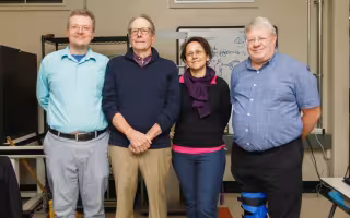 three men and a woman stand in a row facing the camera in front of a whiteboard scrawled with figures and shapes
