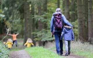 A woman with gray hair walks down a woodland path with her arm around a young child walking beside her; both are wearing purple jackets. Three children run ahead on the trail; two are wearing yellow jackets, one is in an orange T-shirt. 
