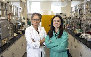 A older woman with short light brown hair and glasses in a white lab coat stands with a younger woman with long dark hair, glasses and a green lab coat in a chemistry laboratory with tools, beakers and chemicals.