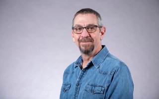 A man with short gray hair, a goatee and glasses wears a denim jacket and poses for a portrait against a gray backdrop.