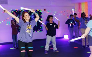 Children and adults wave glow sticks in a room that is lit purple and decorated with glow-in-the-dark stars and tape.