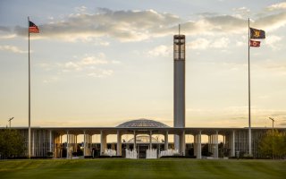 The UAlbany Academic Podium, Carillon and entrance fountain are seen at dusk under a darkening sky. Flags fly on poles on either side of the image.