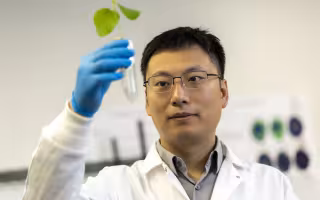 A man with short black hair and glasses in a white lab coat and blue gloves holds up a vial containing a plant