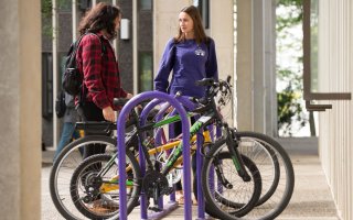 Two women talk while standing next to bikes in a purple bike rack.
