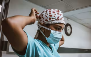 A person wearing light blue medical scrubs and a surgical cap secures a paper face mask around their head. 