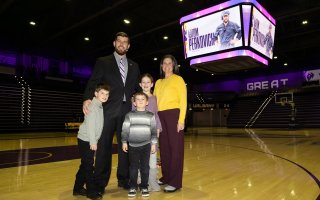A tall man in a dark suit stands with a woman and three children on a basketball court with the scoreboard reading "Tom Perkovich" with purple accents.