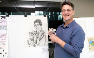 A man wearing glasses and a blue shirt holding a small stuffed mouse toy stands in front of a portrait of himself holding a mouse sketched on paper.