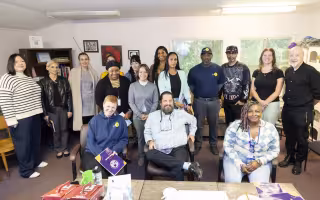 A group of sixteen people pose for a group portrait in a common area at Project Safe Point's Albany location. The room has multiple windows, artwork hung on the walls, and is lined in shelves containing books and bins of personal care supplies.