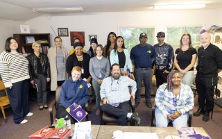 A group of sixteen people pose for a group portrait in a common area at Project Safe Point's Albany location. The room has multiple windows, artwork hung on the walls, and is lined in shelves containing books and bins of personal care supplies.