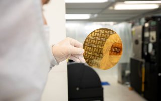 A gloved hand holds a gold-colored silicon wafer reflecting the image of a person in a cleanroom "bunny" suit.