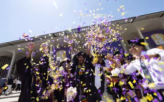 Students in caps and gowns celebrate as purple and gold confetti rain down on them at UAlbany's commencement ceremony.