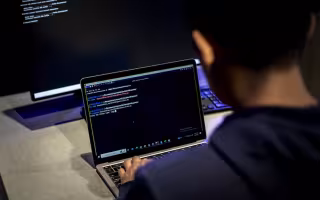 A man sits at a desk with a desktop computer and types code on a laptop.