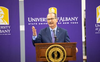 President Rodriguez stands behind a podium in front of UAlbany banners
