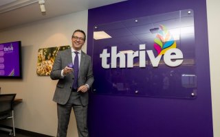 A man in a gray suit smiles while holding up a purple heart in front of a purple wall that says "thrive" in white letters.