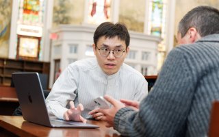 A man with dark hair and glasses sits at a table with a laptop in front of him and conversing with another man at the table holding a cellphone inside a picturesque library.