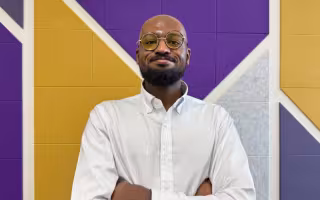 A man wearing yellow tinted glasses and a white, pinstriped button-down shirt poses for a portrait, hands folded across his chest, in front of a wall covered with decorative gold, purple and gray panels.