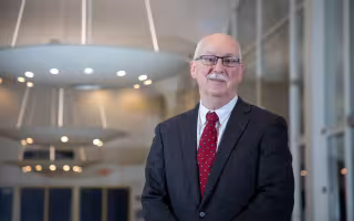 A bald man wears a red tie and a dark suit while standing in front of lighting fixtures hanging from the ceiling indoors.