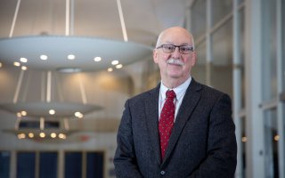 A bald man wears a red tie and a dark suit while standing in front of lighting fixtures hanging from the ceiling indoors.