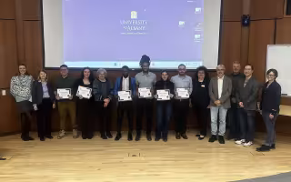 Fourteen people pose for a group portrait in front of a pull-down projection screen displaying the UAlbany logo. All are smiling and six are holding white paper certificates.