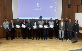 Fourteen people pose for a group portrait in front of a pull-down projection screen displaying the UAlbany logo. All are smiling and six are holding white paper certificates.