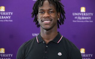 An African Americna young man wears a black collared shirt while standing against a purple backdrop with University at Albany written on it.