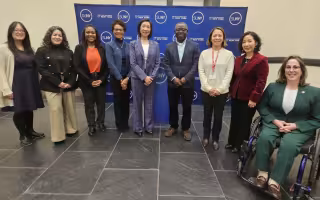 A group of University administrators, faculty and staff pose in front of a blue banner with the words SUNY and State University of New York repeated in white letters.
