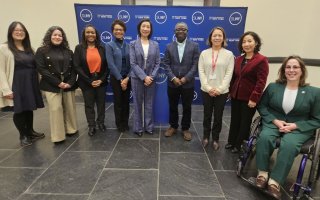 A group of University administrators, faculty and staff pose in front of a blue banner with the words SUNY and State University of New York repeated in white letters.