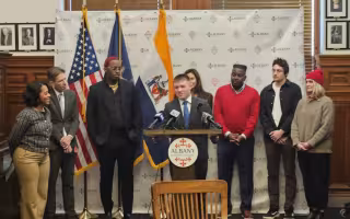 A young man in a suit speaks at a lecturn with a round plaque reading ALBANY on it, as 7 others gather around watching him