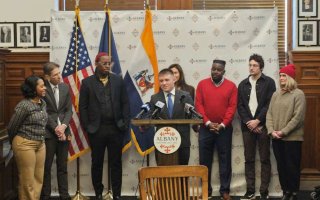 A young man in a suit speaks at a lecturn with a round plaque reading ALBANY on it, as 7 others gather around watching him