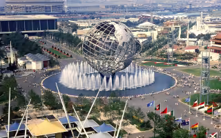 An Elevated view of the 1964 New York World’s Fair in Flushing Meadows with the Unisphere at center and surrounding fairgrounds, buildings and water features visible.