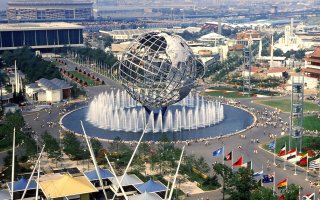 An Elevated view of the 1964 New York World’s Fair in Flushing Meadows with the Unisphere at center and surrounding fairgrounds, buildings and water features visible.