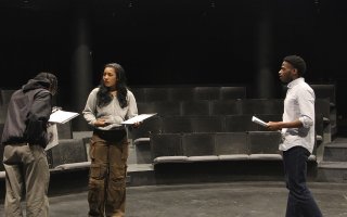 Three students in a darkened theatre hold binders as they practice a production in a theatre-in-the-round room.