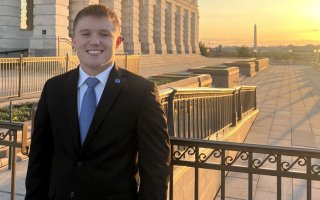 A smiling young man in a suit stands near a marble building with the Washington Monument far in the background.