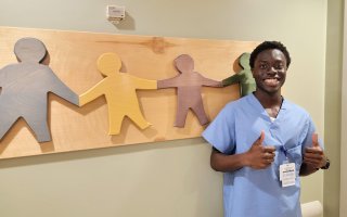 An African American young man in scrubs stands in front of a sign denoting interlinked hands.