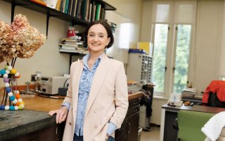 A woman with short brown hair wearing a pale pink blazer and ruffled powder-blue blouse smiles for a portrait in her office.