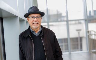 A portrait of a man with eyeglasses in a dark gray hat, black sweater and black jacket leaning against a wall in atrium filled with natural light.