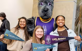 Three students stand with UAlbany's mascot holding UAlbany and Columbia-Greene pennants 