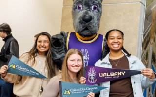 Three students stand with UAlbany's mascot holding UAlbany and Columbia-Greene pennants 