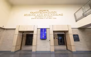 A sign for the College of Nanotechnology, Science, and Engineering hangs above two doorways in a building with light tan and brick walls.