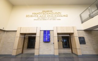 A sign for the College of Nanotechnology, Science, and Engineering hangs above two doorways in a building with light tan and brick walls.