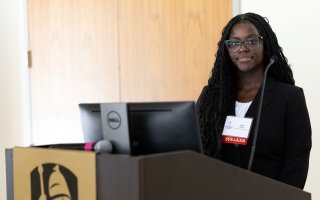 A young African American woman stands at a podium with a gold emblem with the UAlbany logo smiling with a badge that says "speaker"